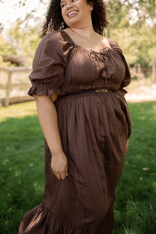 A woman wearing the Ivy City Co Maleah Dress in Warm Cocoa, featuring puff sleeves and a maxi length, stands smiling on the grass with trees and a fence in the background.