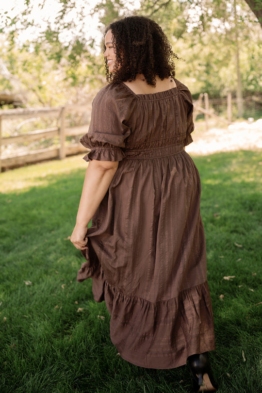 A person with curly hair wears the Ivy City Co Maleah Dress in Warm Cocoa, walking on green grass in a sunlit outdoor setting with trees and a wooden fence in the background.