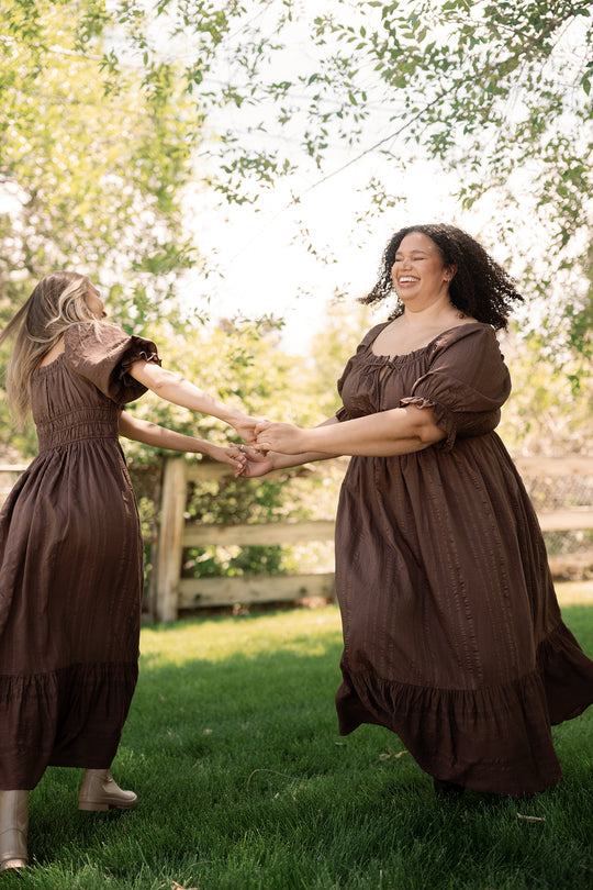 Two women in matching Ivy City Co Maleah Dresses in Warm Cocoa laugh and hold hands while spinning in a grassy outdoor area with sunlight and trees in the background.