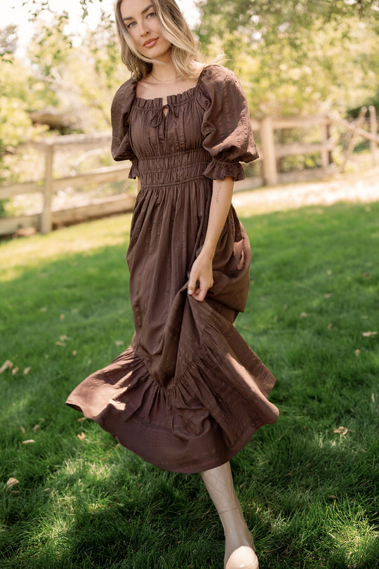 A woman stands on grass by a wooden fence and sunlit trees, gently holding her skirt in the Ivy City Co Maleah Dress in Warm Cocoa. The maternity-friendly dress, featuring puff sleeves, flatters her figure as she poses. She wears cream boots and has long blonde hair.