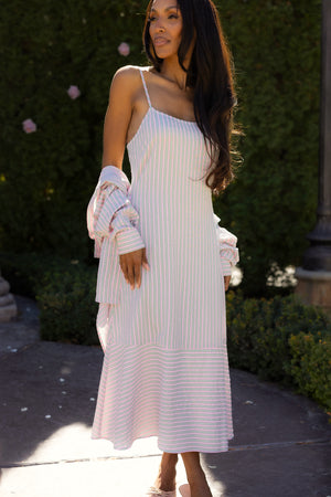 A woman with long hair stands outdoors in sunlight and greenery, wearing the Marbella Dress in Green & Pink Pinstripe by Ivy City Co. She holds a matching long-sleeve shirt draped over her arms.