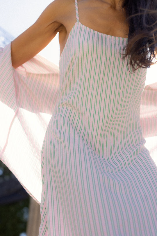 A woman in the Ivy City Co Marbella Dress in Green & Pink Pinstripe stands outdoors, sunlight highlighting the fabric as she raises her arms, showcasing the dress’s lightness and flow.