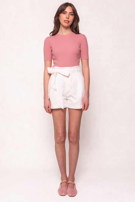 A woman stands facing forward, wearing a short-sleeved pink ribbed top, Ivy City Co’s Maria Shorts in White Eyelet with a removable tie, and pink flats. The background is plain and light-colored.