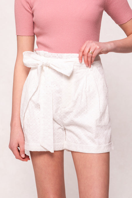 A person wears Ivy City Co’s Maria Shorts in White Eyelet, featuring a high waist and removable tie, paired with a pink ribbed short-sleeve top against a plain light background.