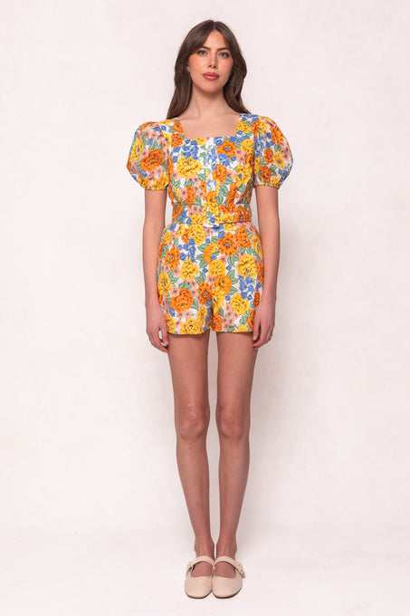 A woman stands facing forward in the Marianne Romper in Floral by Ivy City Co, featuring puffed short sleeves and beige flats. This stylish cotton romper is set against a plain, light-colored background.