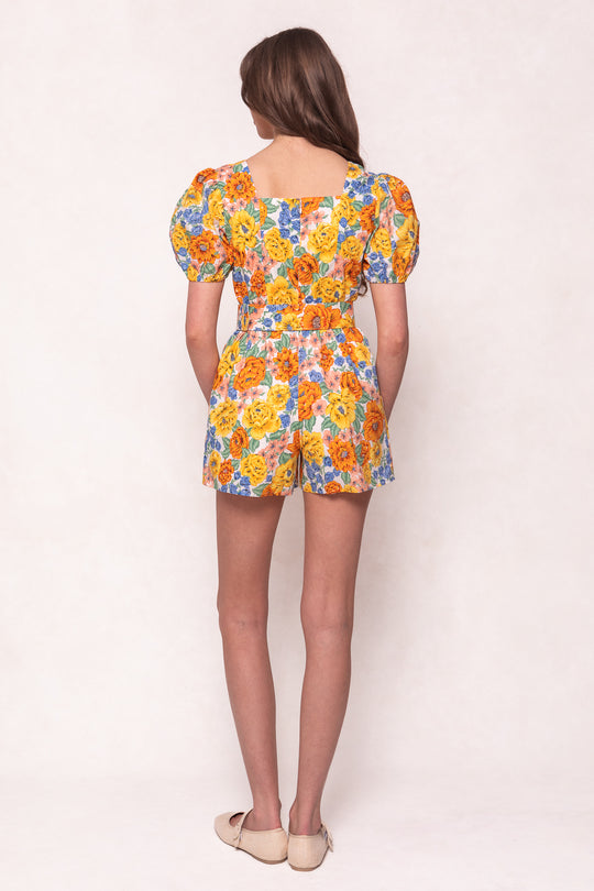 A woman with long hair stands facing away in the Marianne Romper in Floral by Ivy City Co, featuring short puff sleeves and a square neckline. She wears beige flats and is set against a plain, light background.