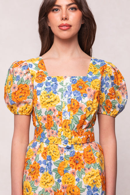 A woman wears the Marianne Romper in Floral by Ivy City Co, featuring puffed short sleeves, a square neckline, vibrant yellow, orange and blue flowers with green leaves, front buttons for nursing, and a matching waist belt.