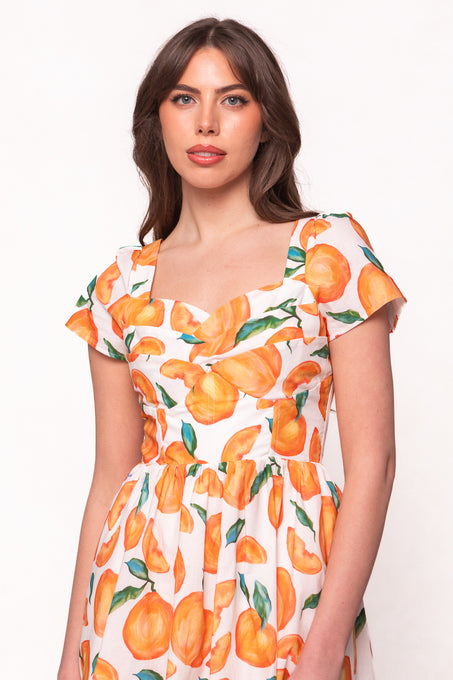 A woman with long brown hair wears the Marie Dress in Citrus by Ivy City Co—a white dress with a peach fruit print, tiered midi skirt, short sleeves, and sweetheart neckline—standing before a plain background.
