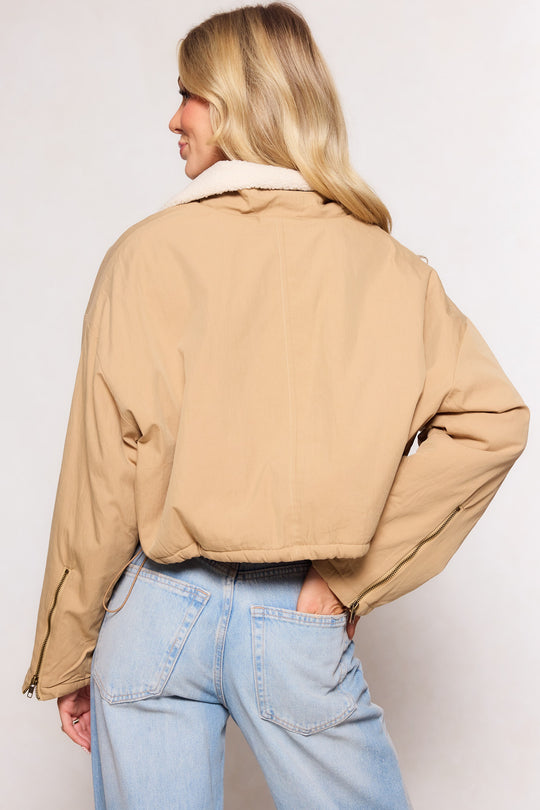 A woman with long blonde hair wears the Ivy City Co Marleigh Jacket in Tan, featuring a white fleece collar, paired with light blue high-waisted jeans. She stands with her back to the camera and one hand in her pocket.