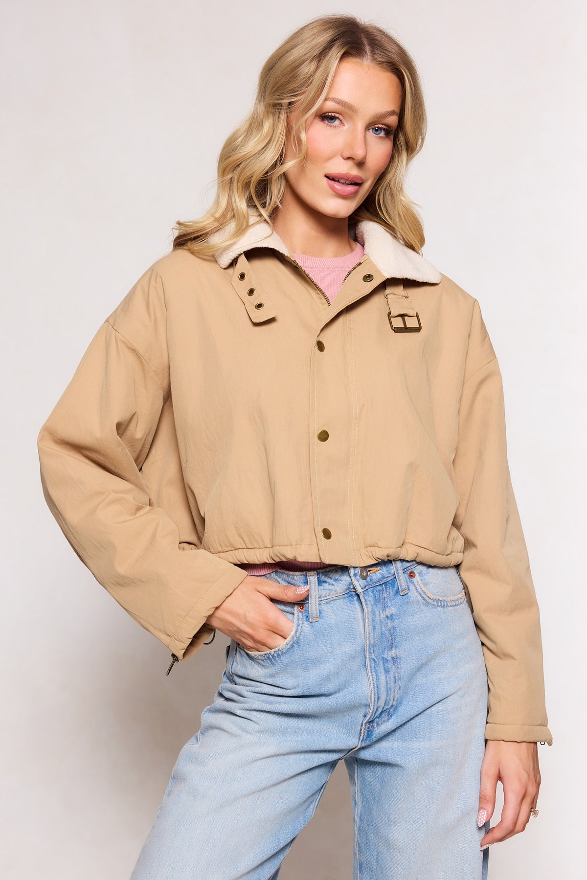 A woman poses with one hand in her pocket, wearing the Ivy City Co Marleigh Jacket in Tan over a light pink top and light blue high-waisted jeans, set against a plain background.