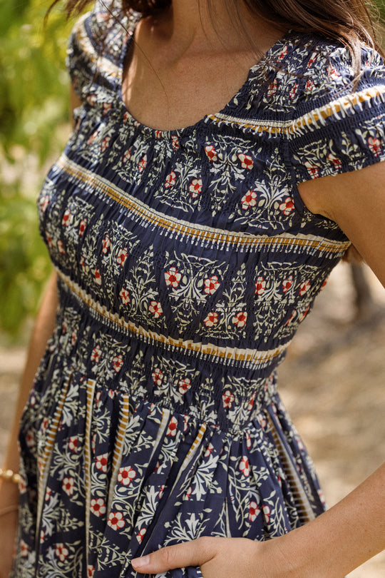 A woman wearing the Ivy City Co Marlow Cap Sleeve Dress in Navy Fleur stands outdoors. Her face is not shown; only her torso and arm are visible, drawing attention to the smocked bodice of this maternity nursing-friendly style. The background features softly blurred greenery.