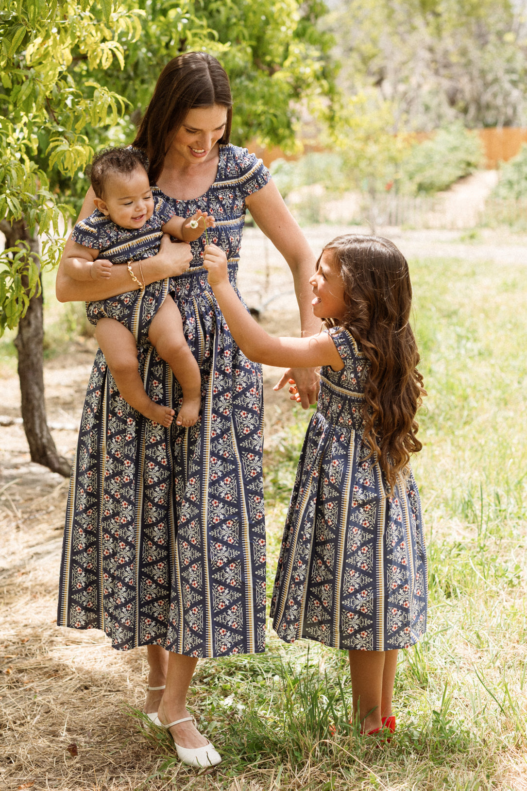 A woman and two young girls, all dressed in the Ivy City Co Marlow Cap Sleeve Dress in Navy Fleur, stand outdoors in a sunny, grassy area with trees. The woman’s maternity and nursing-friendly dress features a smocked bodice as she holds a baby, while the older girl reaches up to her.