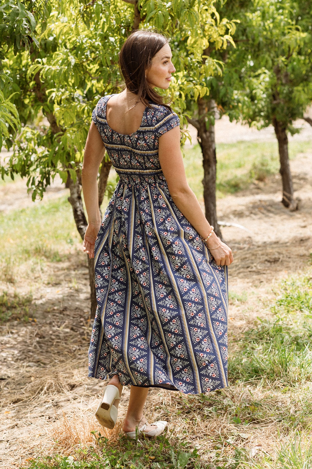 Wearing the Ivy City Co Marlow Cap Sleeve Dress in Navy Fleur, a woman stands outdoors on the grass, holding her skirt and looking over her shoulder. The dress features a blue patterned design with a smocked bodice, making it both stylish and maternity nursing friendly. She is surrounded by green trees and sunlight.