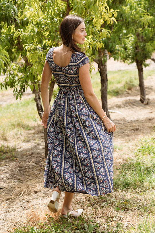 Wearing the Ivy City Co Marlow Cap Sleeve Dress in Navy Fleur, a woman stands outdoors on the grass, holding her skirt and looking over her shoulder. The dress features a blue patterned design with a smocked bodice, making it both stylish and maternity nursing friendly. She is surrounded by green trees and sunlight.