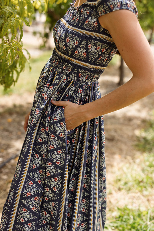A person wears the Ivy City Co Marlow Cap Sleeve Dress in Navy Fleur—a short-sleeved maxi dress with a smocked bodice and a floral and striped pattern—while standing outdoors with one hand in their pocket, surrounded by greenery and sunlight.