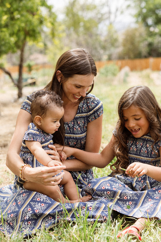 A woman and two young girls sit on the grass outdoors, all wearing matching Ivy City Co Marlow Cap Sleeve Dresses in Navy Fleur. They are smiling and playing together in a sunlit, natural setting.