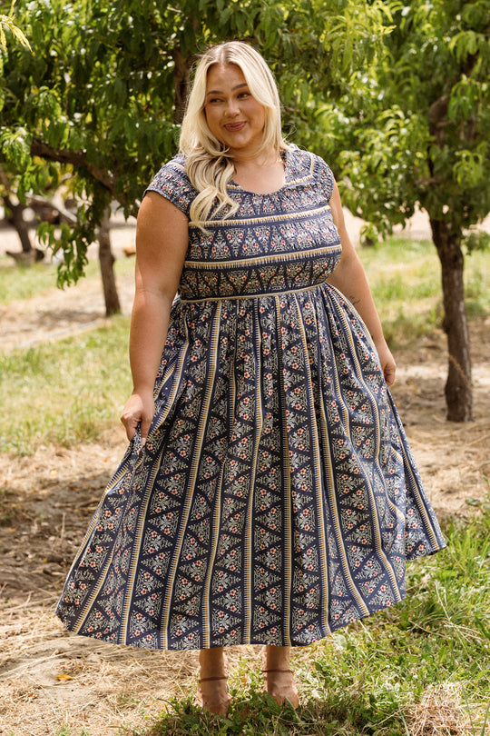 A woman with long blonde hair stands in a sunny orchard, smiling at the camera as she holds the skirt of her Ivy City Co Marlow Cap Sleeve Dress in Navy Fleur. The patterned blue and white dress features a smocked bodice that adds charm while making it both maternity and nursing friendly.