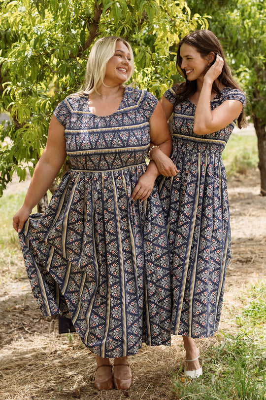 Two women stand arm in arm outdoors beneath leafy trees on a sunny day, smiling at each other while wearing matching Ivy City Co Marlow Cap Sleeve Dresses in Navy Fleur—stylish outfits designed to be both maternity and nursing friendly.