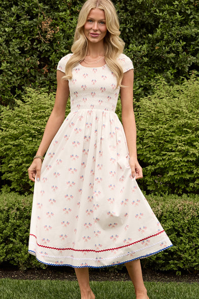A woman stands on grass before green bushes, smiling and holding her skirt. She wears the Ivy City Co Marlow Cap Sleeve Dress in Star Spangled, an ankle-length, maternity-friendly dress with a blue and red star print and trim at the hem.