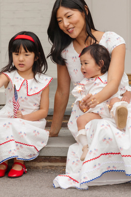 A woman and two young girls, all wearing Ivy City Co’s Marlow Cap Sleeve Dress in Star Spangled, sit on steps with festive Fourth of July accessories. The dress features red and blue trim and is even maternity friendly!.
