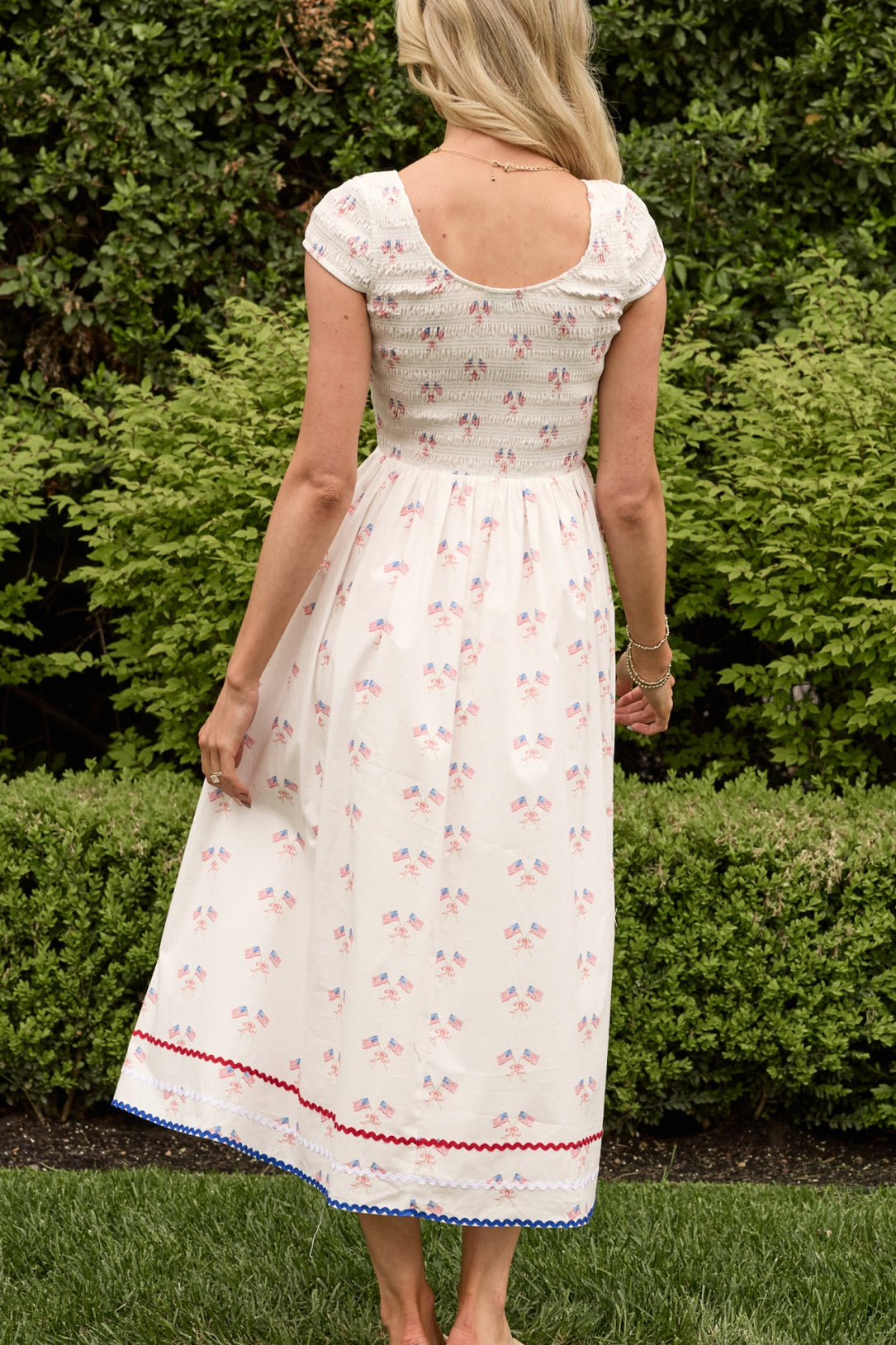 A woman with long blonde hair stands barefoot on grass, facing away, wearing the Ivy City Co Marlow Cap Sleeve Dress in Star Spangled, a maternity-friendly Fourth of July dress; lush green bushes are in the background.