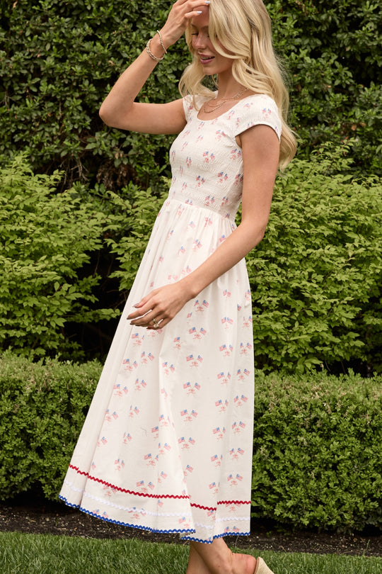 A woman with long blonde hair smiles and touches her hair while standing outdoors in front of green shrubs, wearing the Ivy City Co Marlow Cap Sleeve Dress in Star Spangled.