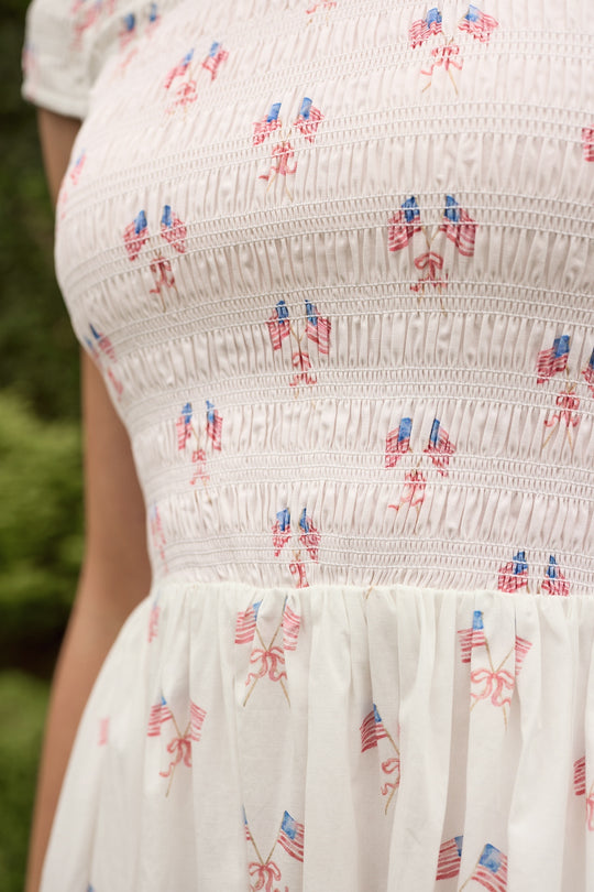 A close-up of the Ivy City Co Marlow Cap Sleeve Dress in Star Spangled—a white, maternity-friendly dress featuring smocked details and a colorful star print. The dress is worn outdoors with blurred greenery in the background.