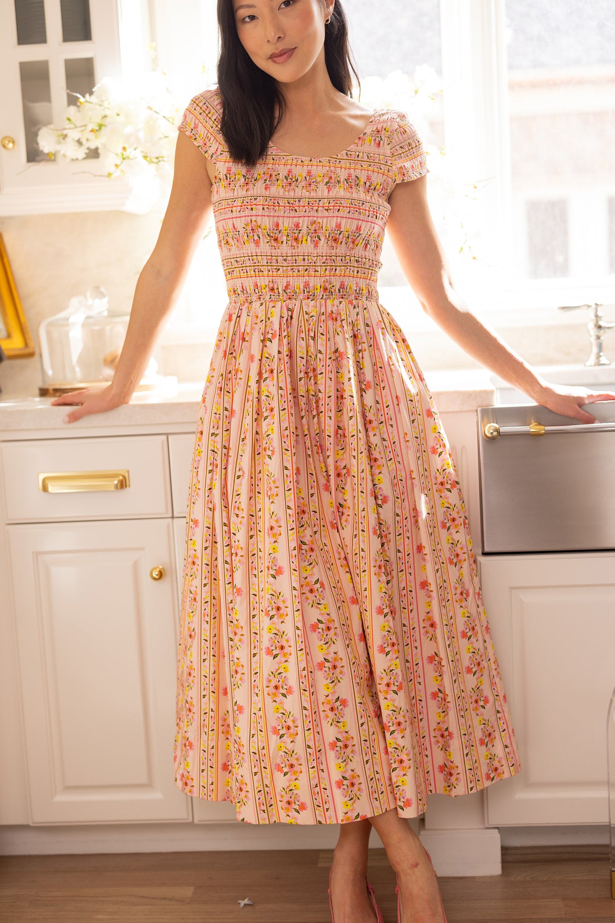 A woman in the Ivy City Co Marlow Cap Sleeve Dress in Peach Handpainted Flora Stripe stands in a bright kitchen with white cabinets and a farmhouse sink—a lovely, spring-ready maternity-friendly portrait dress.