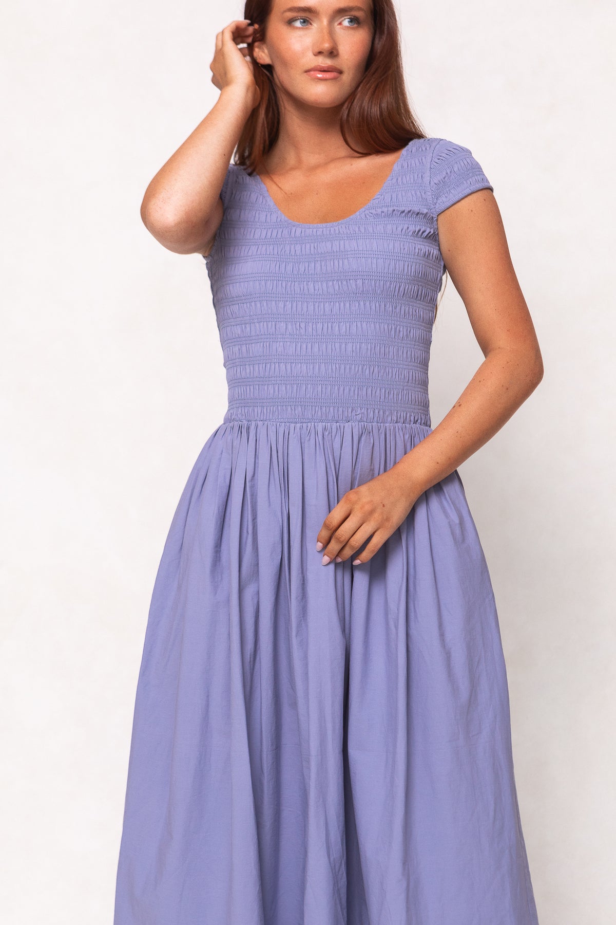 A woman wearing the Ivy City Co Marlow Cap Sleeve Dress in Blue, featuring a fitted bodice and flowing skirt, stands against a plain light background, touching her hair with one hand.