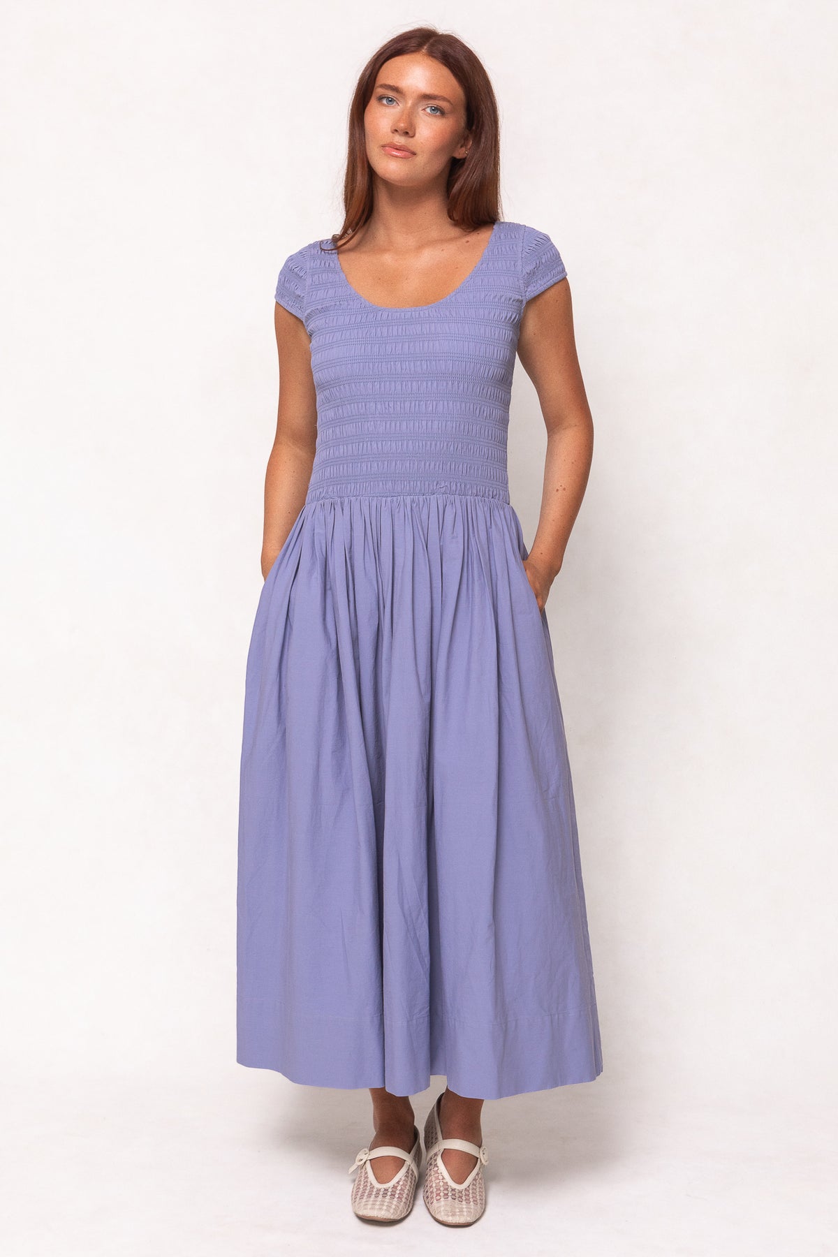 A woman with long brown hair wears the Ivy City Co Marlow Cap Sleeve Dress in Blue, an ankle-length, nursing-friendly dress with a smocked bodice and pockets, paired with white woven flats, against a plain light background.