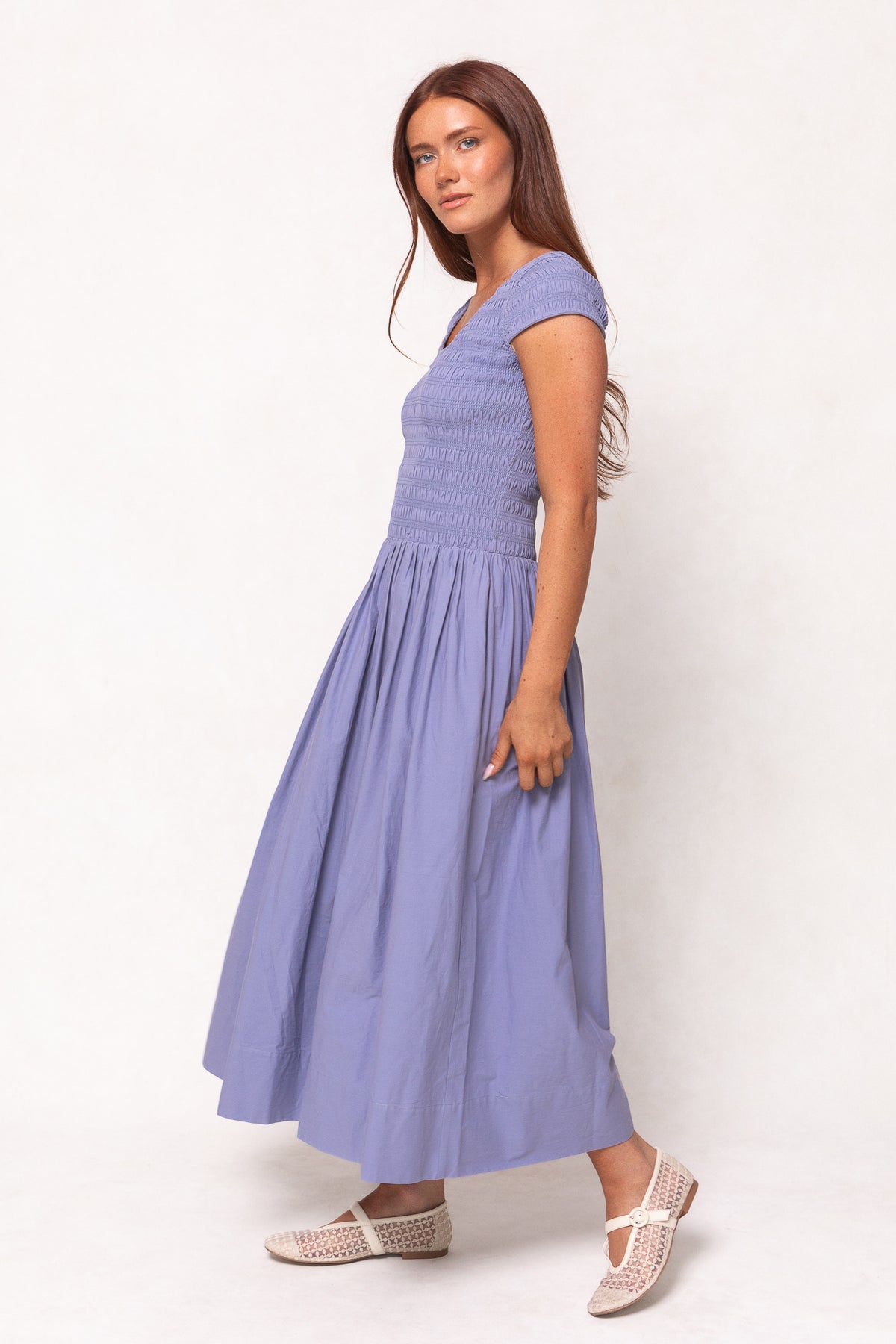 A woman with long brown hair stands sideways against a plain light background, wearing the Ivy City Co Marlow Cap Sleeve Dress in Blue and white woven flats.