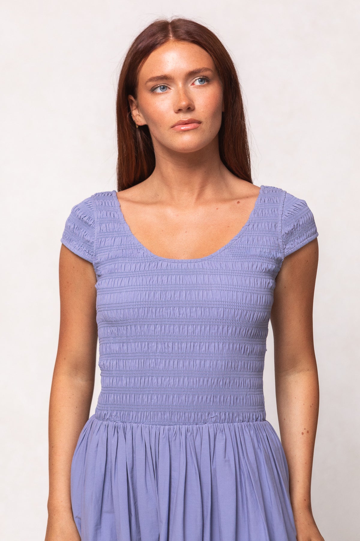 A woman with long reddish-brown hair wears the Ivy City Co Marlow Cap Sleeve Dress in Blue, featuring a nursing-friendly design with short sleeves and a smocked bodice, as she looks slightly to the side against a plain light background.