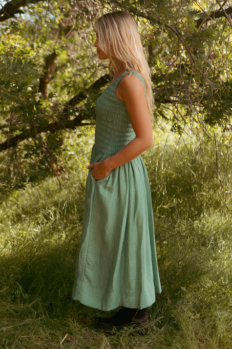 A woman with long blonde hair stands sideways in a grassy area under tree branches, wearing the Ivy City Co Marlow Dress in Green—maternity friendly with a smocked bodice and handy pockets.