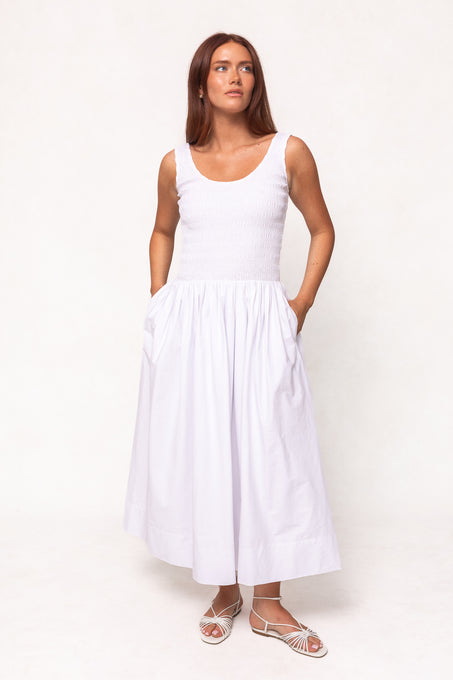 A woman with long brown hair stands against a light background, looking to the side with hands in her pockets, wearing Ivy City Co’s Marlow Dress in White—a cotton midi dress with fitted bodice and full skirt, nursing friendly, paired with white sandals.
