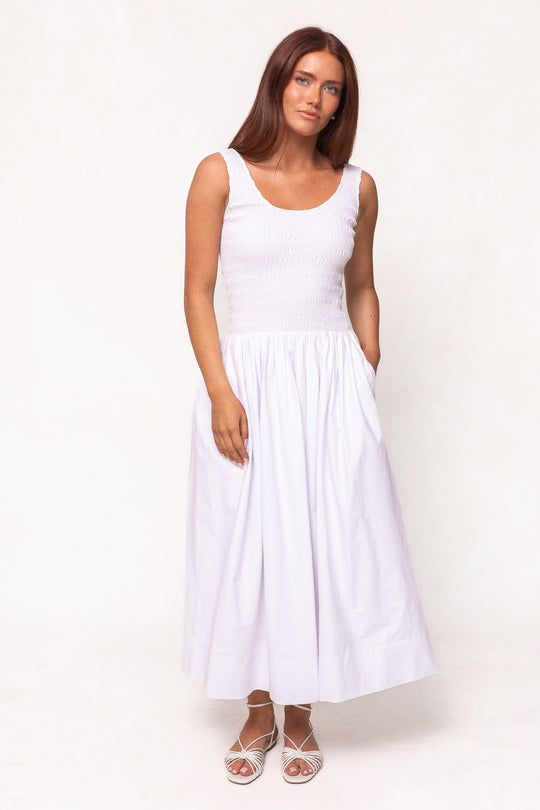 A woman with long brown hair wears the Ivy City Co Marlow Dress in White, a sleeveless cotton midi featuring a fitted bodice and gathered skirt. Maternity friendly, she stands facing the camera on a plain white background.