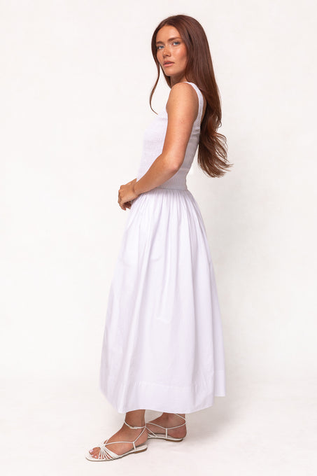 A woman with long reddish-brown hair stands sideways, looking at the camera in the Ivy City Co Marlow Dress in White— a sleeveless cotton midi style paired with strappy white sandals. The look is maternity friendly and effortlessly chic against a light background.