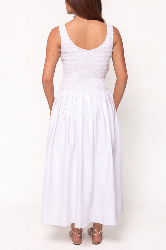 A woman with long brown hair stands facing away, wearing the Ivy City Co Marlow Dress in White—a sleeveless cotton midi dress with a smocked bodice and flowing skirt—paired elegantly with strappy silver heels. Maternity friendly.