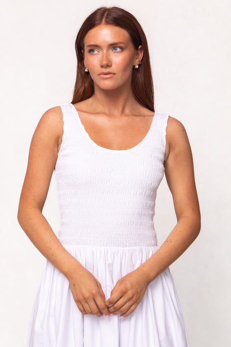 A woman with long brown hair wears the Ivy City Co Marlow Dress in White, a sleeveless cotton midi with a smocked bodice. She stands against a light background, looking slightly to the side. The dress is nursing-friendly and comfortable for maternity.