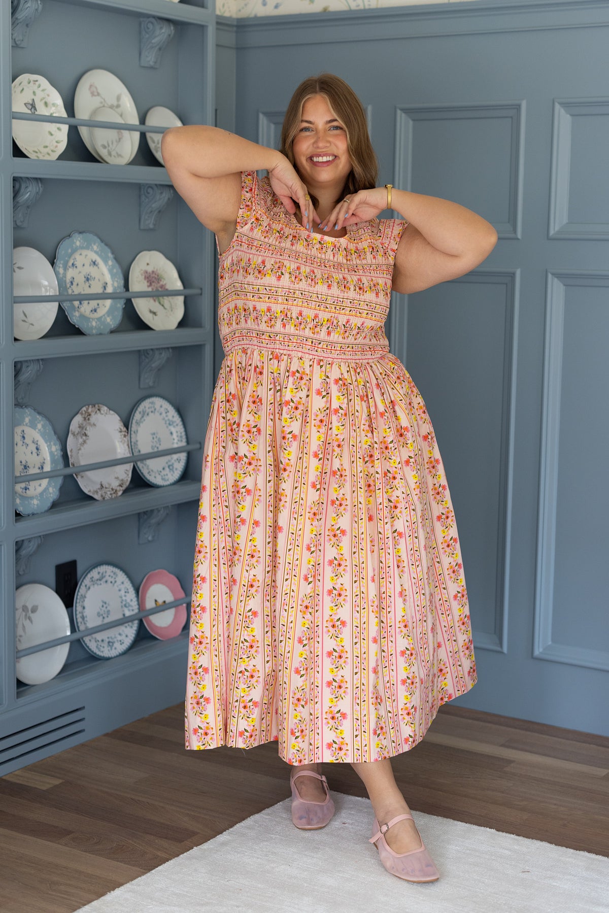 A woman wearing the Marlow Cap Sleeve Dress in Peach Handpainted Flora Stripe by Ivy City Co stands indoors on a wooden floor, smiling in front of a blue wall and shelf with decorative plates, capturing a cheerful spring vibe.