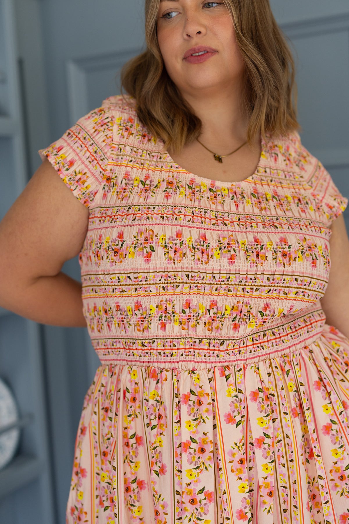 Wearing the Ivy City Co Marlow Cap Sleeve Dress in Peach Handpainted Flora Stripe, a person stands indoors against a blue background, looking slightly to the side—a perfect spring portrait dress that’s also maternity friendly.