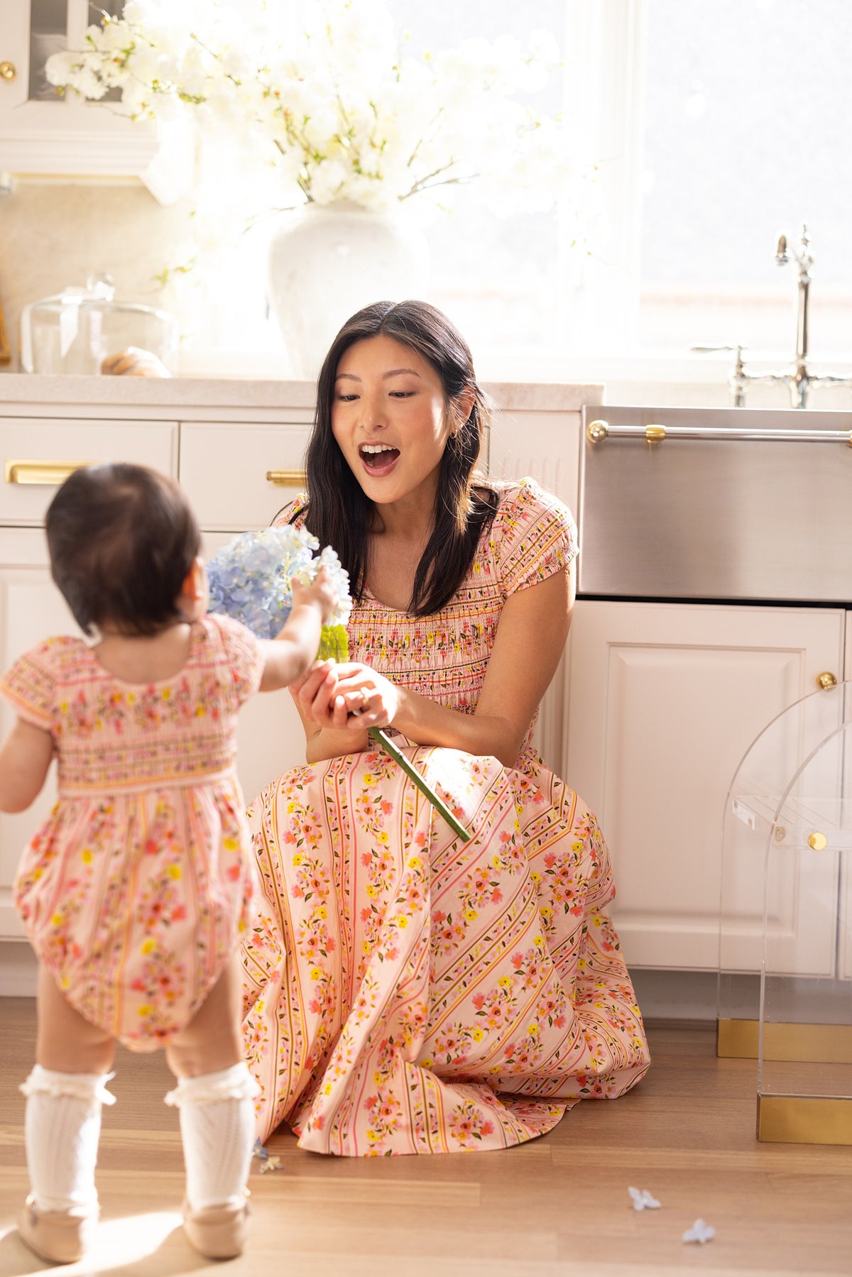 A woman wears the Ivy City Co Marlow Cap Sleeve Dress in Peach Handpainted Flora Stripe, kneeling on the kitchen floor and smiling as a toddler in a matching outfit gives her blue flowers, sunlight streaming in for a warm, cheerful spring scene.
