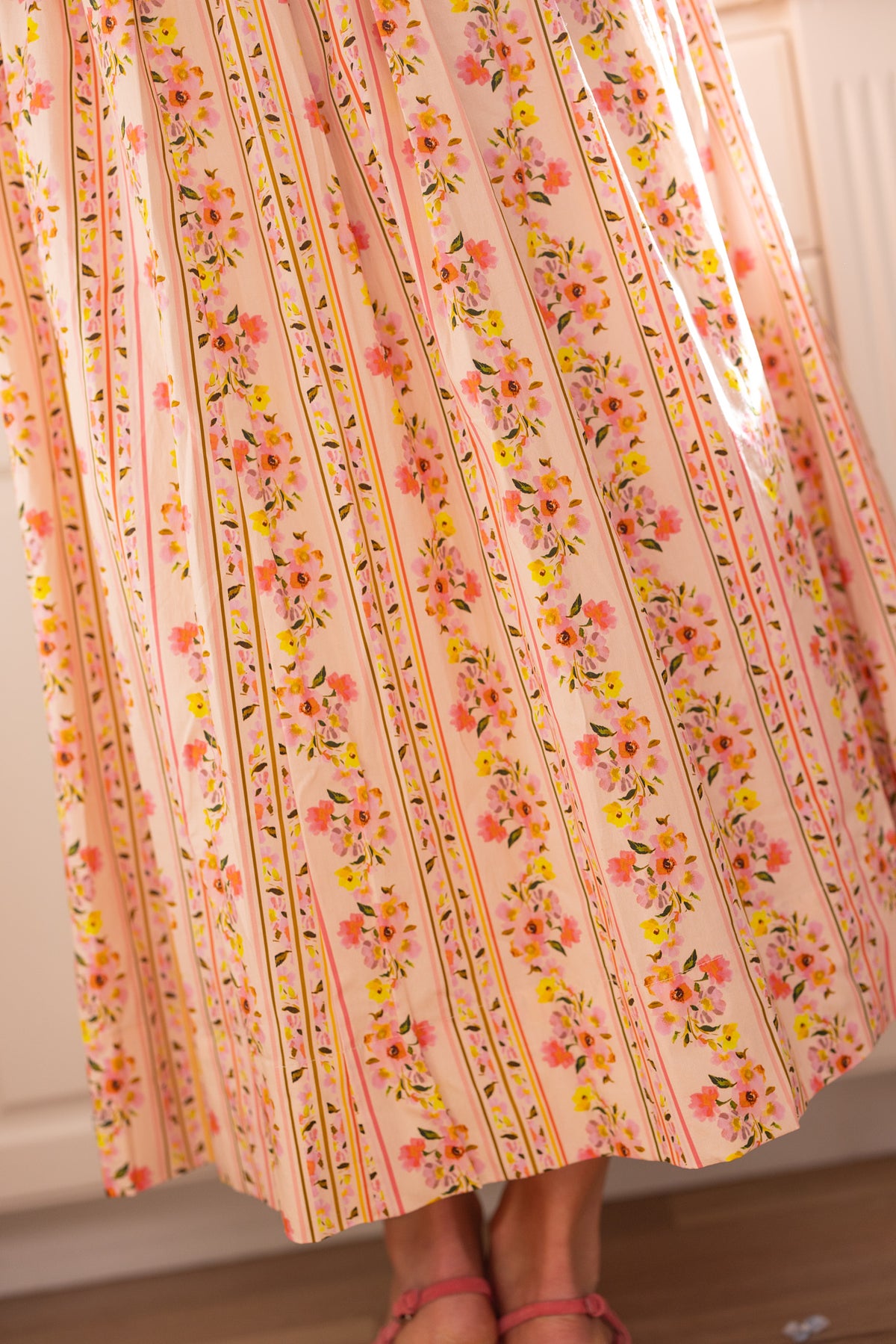 A person stands indoors on a wooden floor, showing only their lower legs and pink sandals, wearing the Ivy City Co Marlow Cap Sleeve Dress in Peach Handpainted Flora Stripe—perfect for spring portraits with its vibrant floral and striped design.