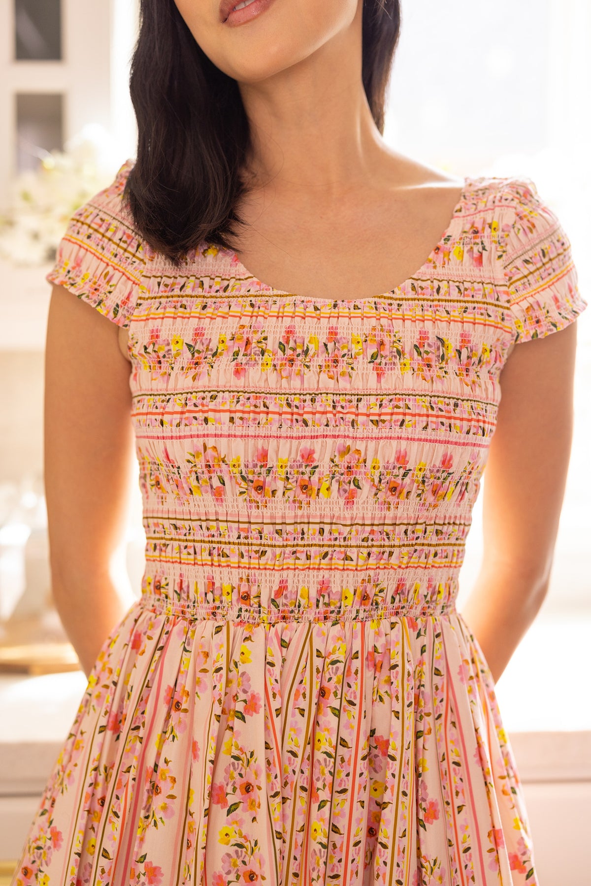 A woman wears the Ivy City Co Marlow Cap Sleeve Dress in Peach Handpainted Flora Stripe, a maternity-friendly spring piece, indoors with soft sunlight. Her face is partially out of frame.