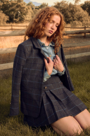 A woman with wavy red hair kneels on grass in the Ivy City Co Marren Blazer in Charcoal Plaid with a matching skirt over a denim shirt. A wooden fence and trees, softly lit by warm sunlight, form the background.