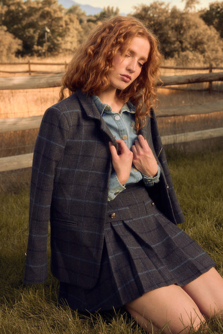 A woman with wavy red hair kneels on grass in the Ivy City Co Marren Blazer in Charcoal Plaid with a matching skirt over a denim shirt. A wooden fence and trees, softly lit by warm sunlight, form the background.
