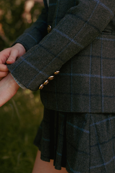 A person adjusts the cuff of the Ivy City Co Marren Blazer in Charcoal Plaid, featuring gold buttons and paired with a matching pleated skirt, outdoors against blurred greenery.