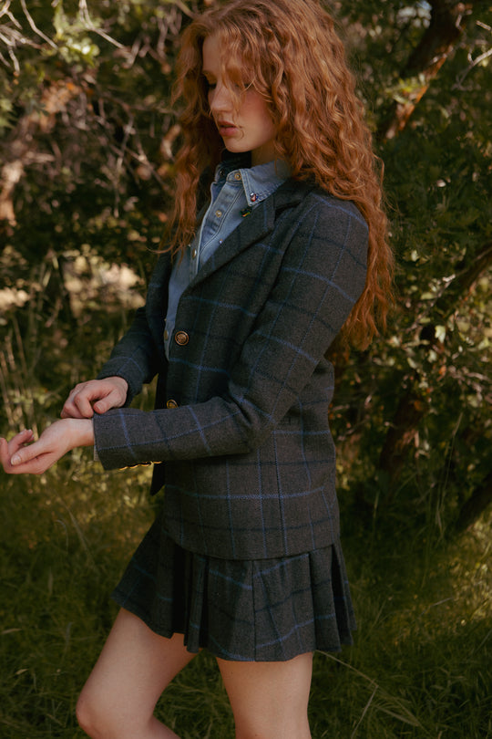 A person with long curly red hair stands outdoors amid greenery, adjusting their sleeves while wearing Ivy City Co’s Marren Blazer in Charcoal Plaid with a matching pleated skirt over a light blue shirt.