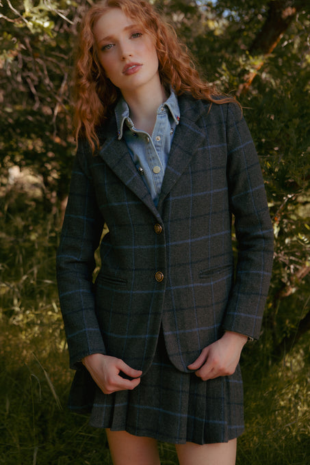 A woman with wavy red hair poses outdoors in the Ivy City Co Marren Blazer in Charcoal Plaid and a pleated skirt over a denim shirt, surrounded by green trees and sunlight.