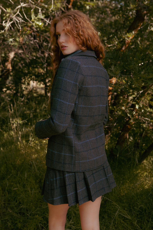 A woman with long, curly red hair stands in a wooded area, wearing the Ivy City Co Marren Blazer in Charcoal Plaid and a matching pleated skirt, glancing over her shoulder at the camera.