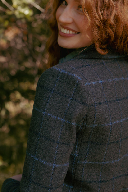 A woman with wavy red hair smiles over her shoulder, wearing the Marren Blazer in Charcoal Plaid by Ivy City Co. A backdrop of blurred greenery completes the scene.